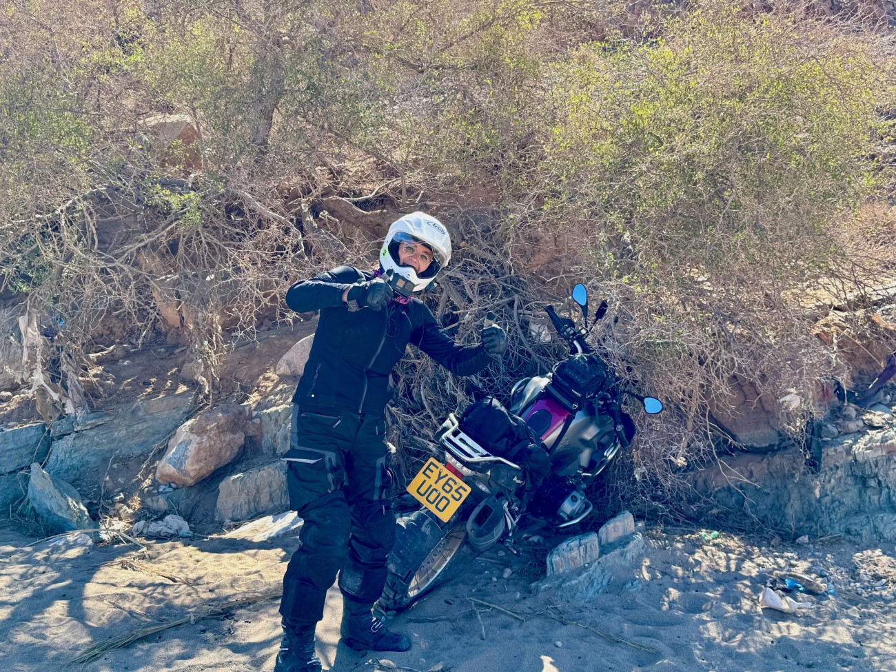 Sophie holding her thumbs up with a motorbike stuck in a bush behind her